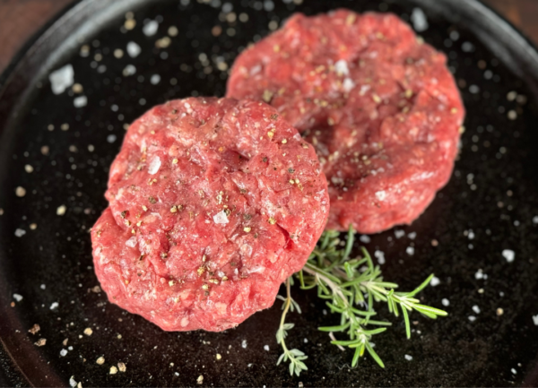 2 Grass fed raw beef patties on cast iron skillet with a sprig of rosemary covered with sea salt and cracked pepper